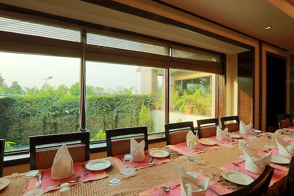 A dining area with a table set for a meal, large windows with blinds, and a view of greenery outside.