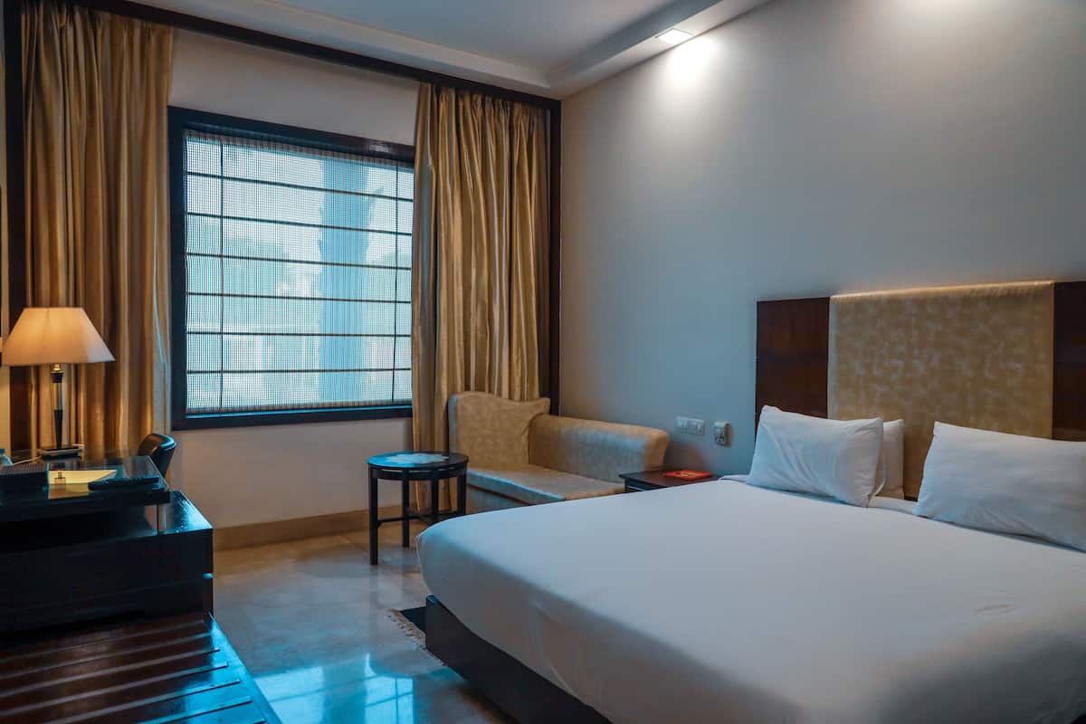 Une chambre d’hôtel avec un lit, un bureau, une chaise, une lampe et une fenêtre avec des rideaux.