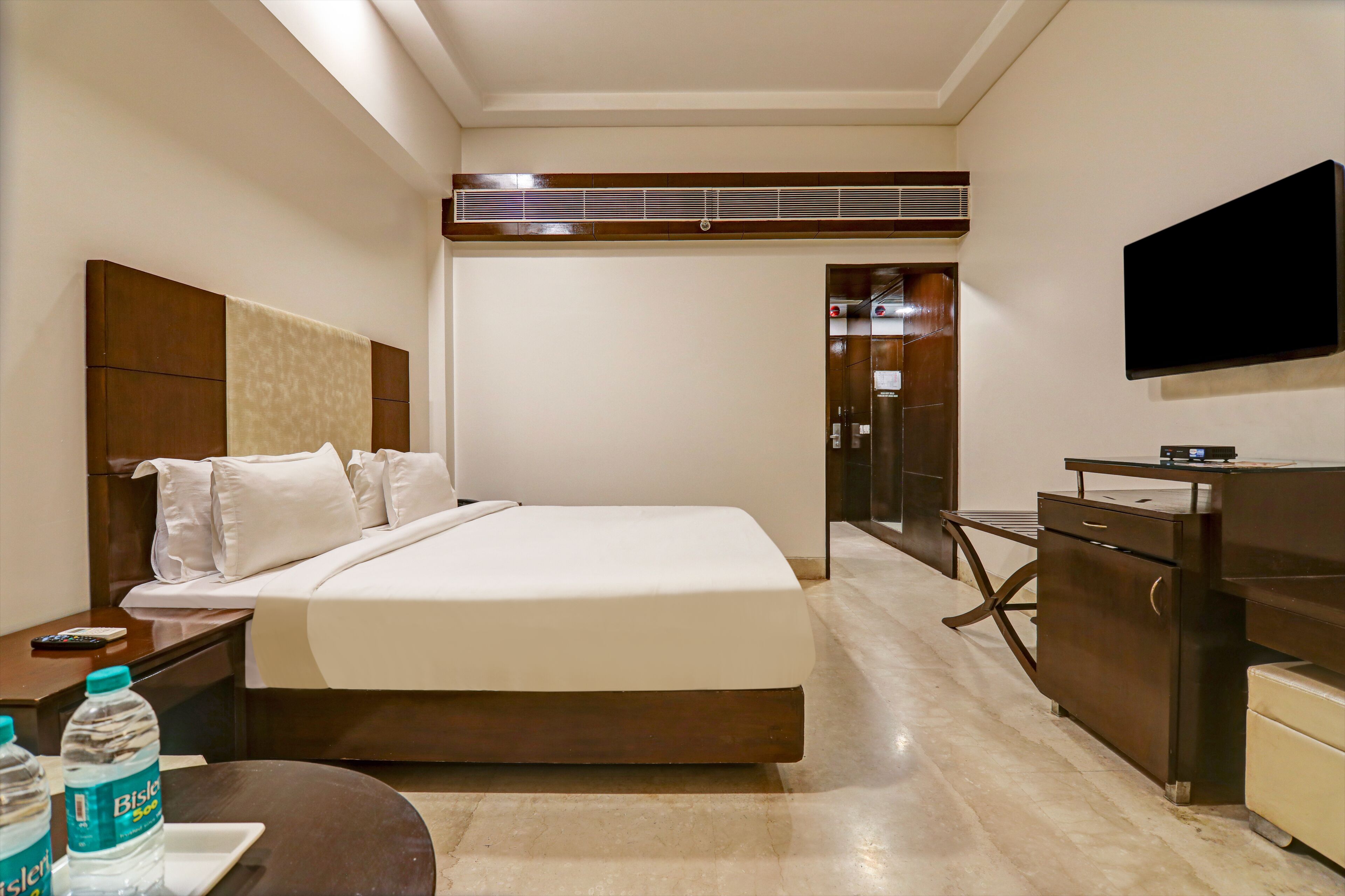 A hotel room with a bed, a TV mounted on the wall, a desk with a chair, and a minibar.