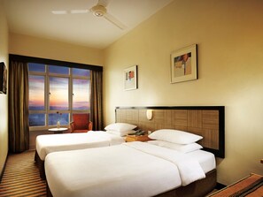 A hotel room with two beds, a window with a view, a ceiling fan, and a framed picture on the wall.