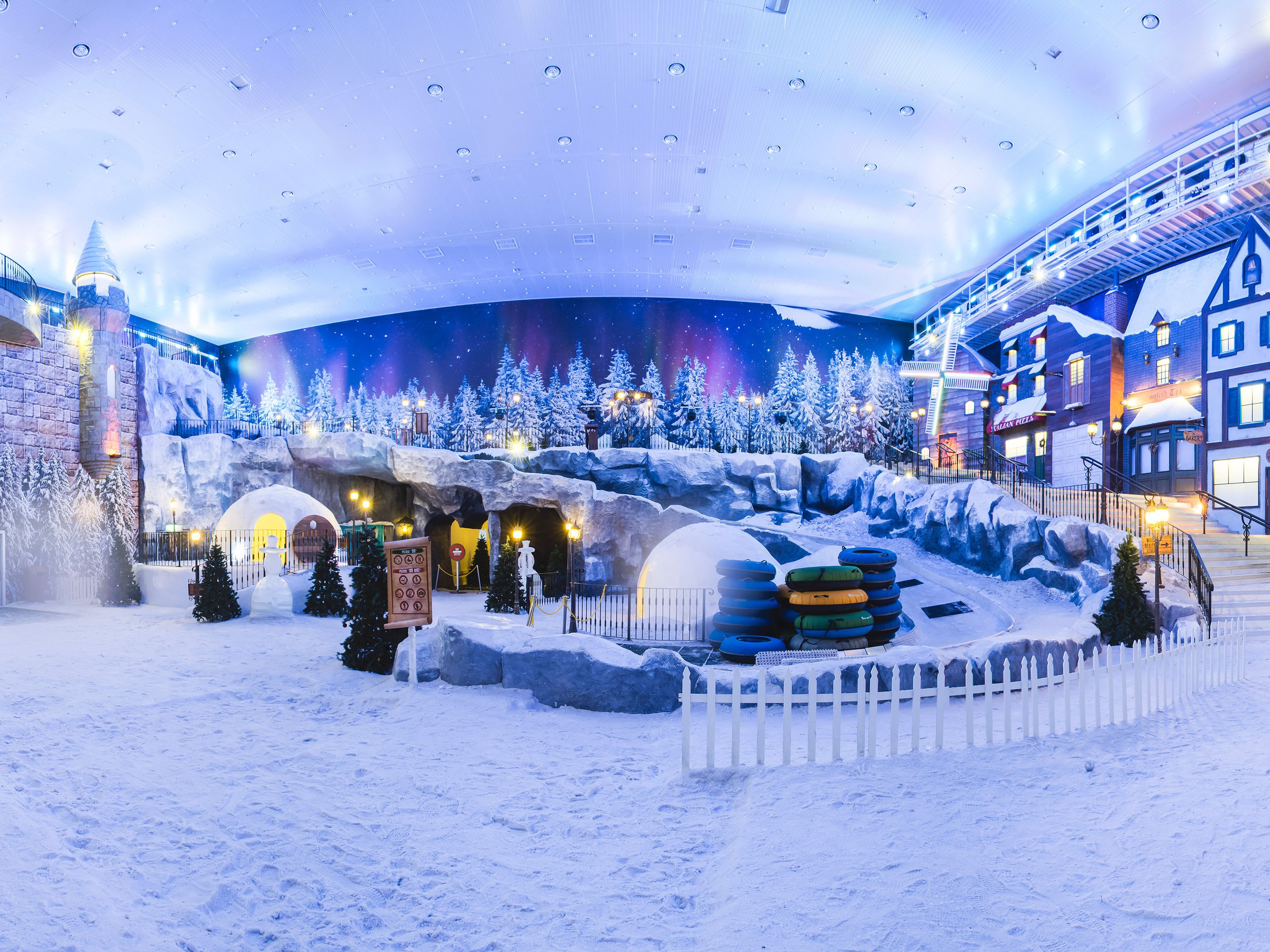 Área externa coberta de neve, com um boneco de neve, uma placa e um cercado branco de piquete.
