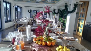 Ein reichhaltiges Buffet mit verschiedenen Speisen, darunter Obst und Gebäck, in einem hell erleuchteten Raum mit großen Fenstern und Zimmerpflanzen.