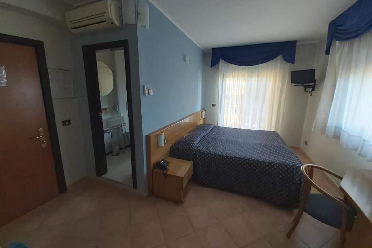 A hotel room with a bed, a chair, a small table, a mirror, an air conditioning unit, and a television.