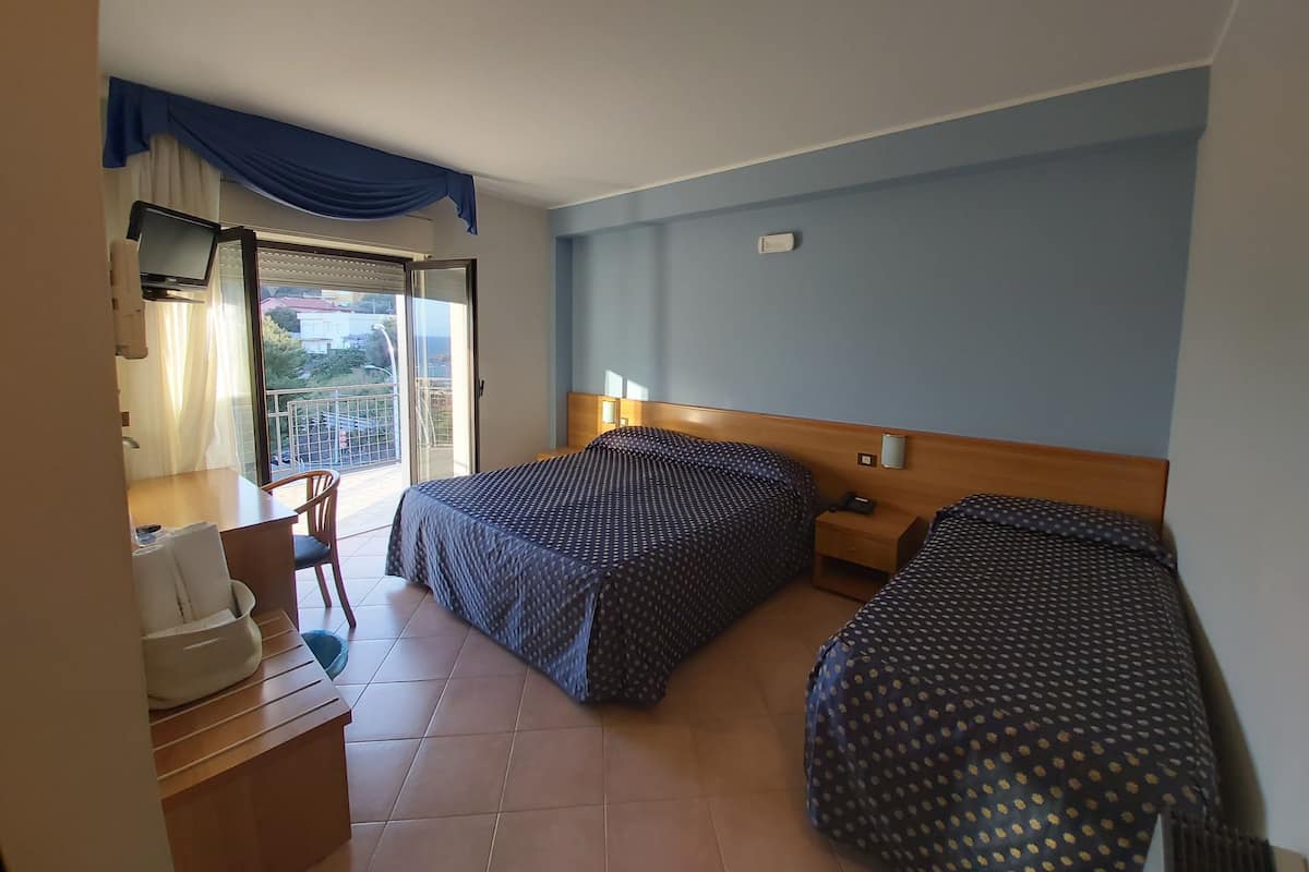 A hotel room with two beds, a desk, a chair, a TV, and a balcony with a view.