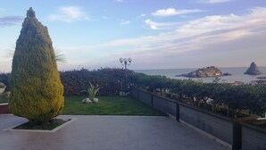 Ein angelegter Garten mit einer hohen Nadelbaumart, einer Laterne und Blick auf das Meer mit Felsformationen.