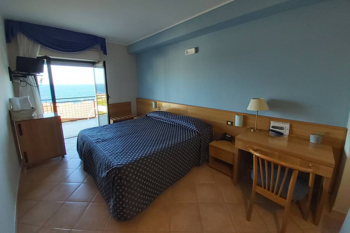 A hotel room with a bed, a desk, a chair, a lamp, a telephone, and a window with a view of the sea.