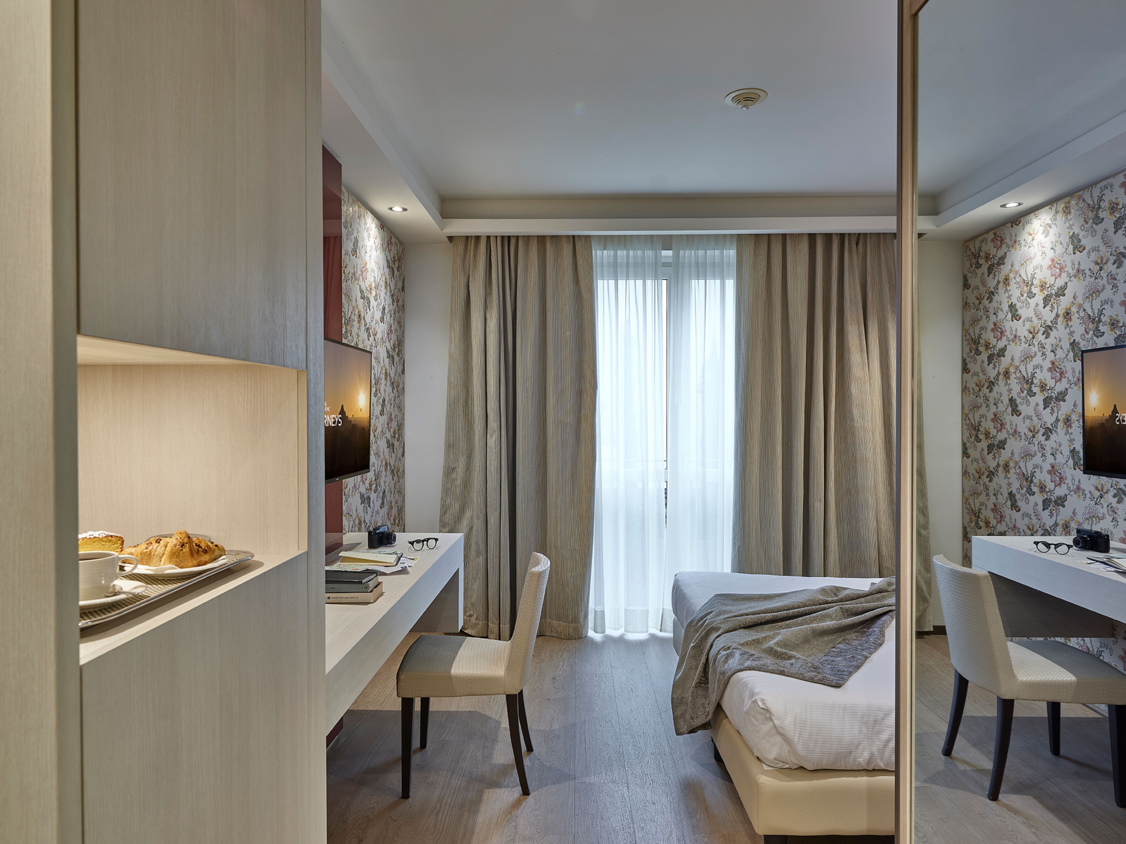 Habitación de hotel con cama, escritorio, silla, televisor y un ventanal con cortinas.