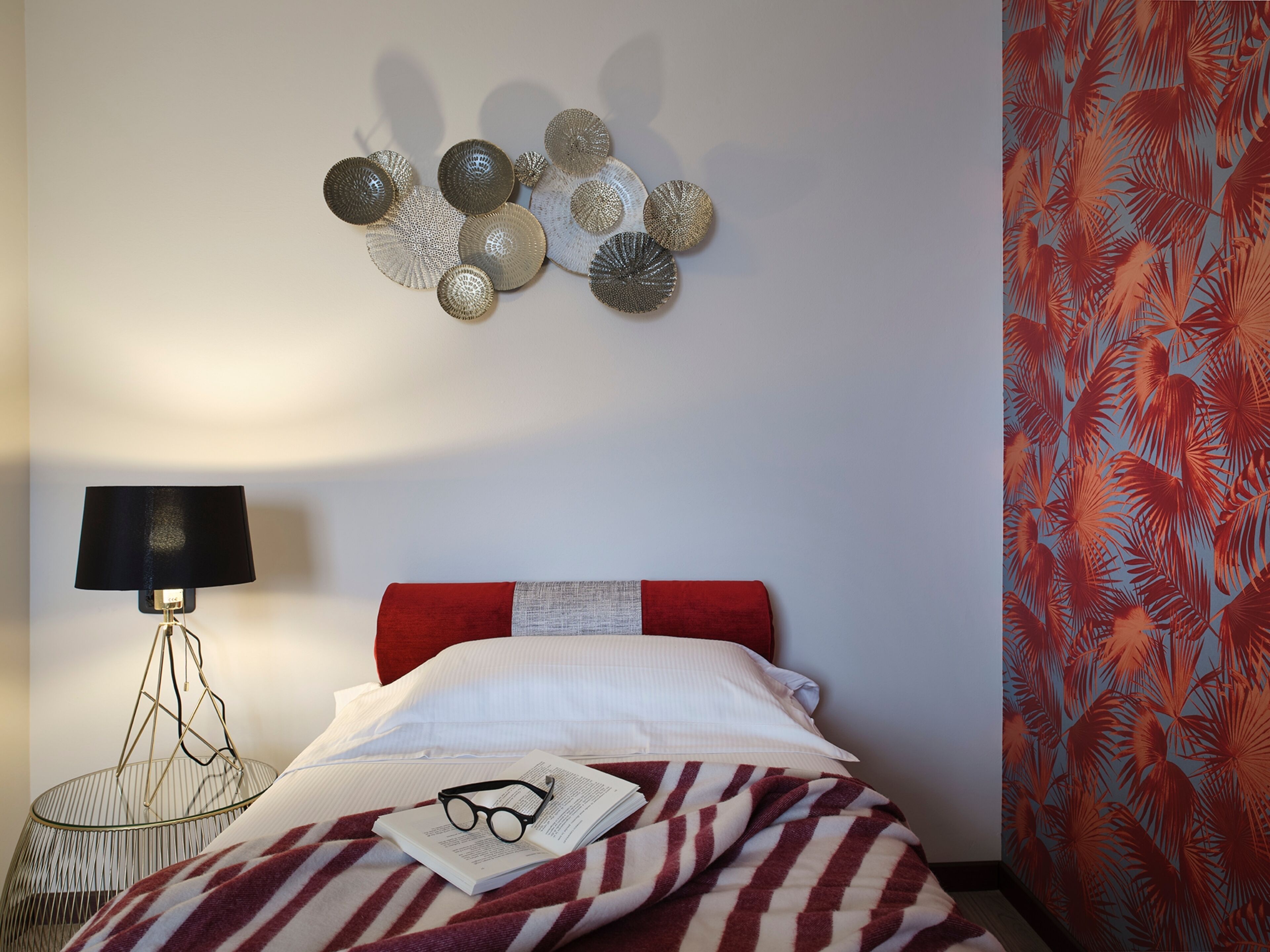 A neatly made bed with a striped blanket, a pair of glasses on a book, a lamp, and decorative wall art.