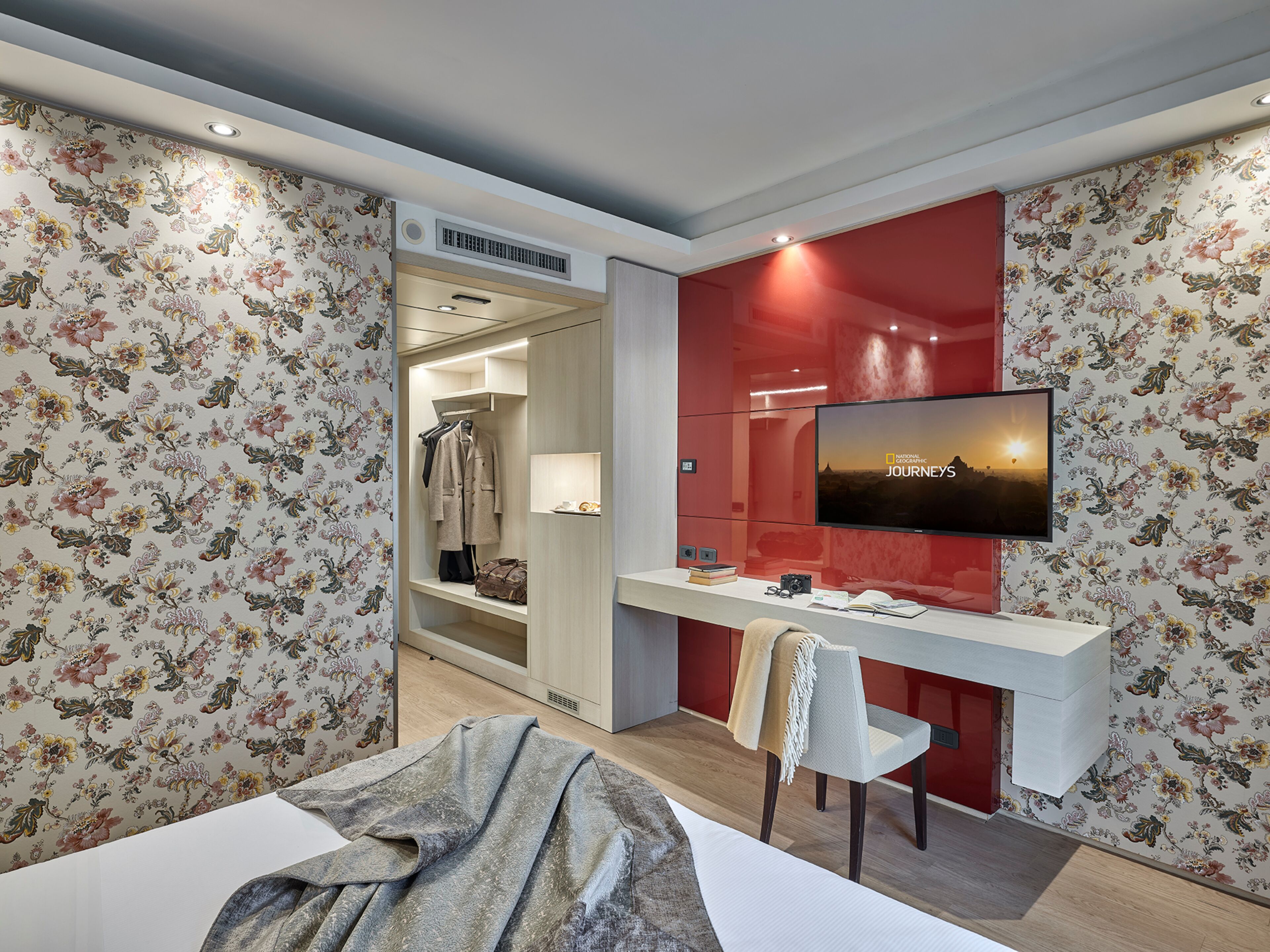 A hotel room with a bed, a desk with a chair, a TV mounted on the wall, and a wardrobe.