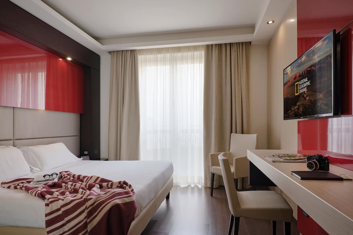 Ein Hotelzimmer mit Bett, Schreibtisch mit Stuhl, Fernseher und einem Fenster mit Vorhängen.