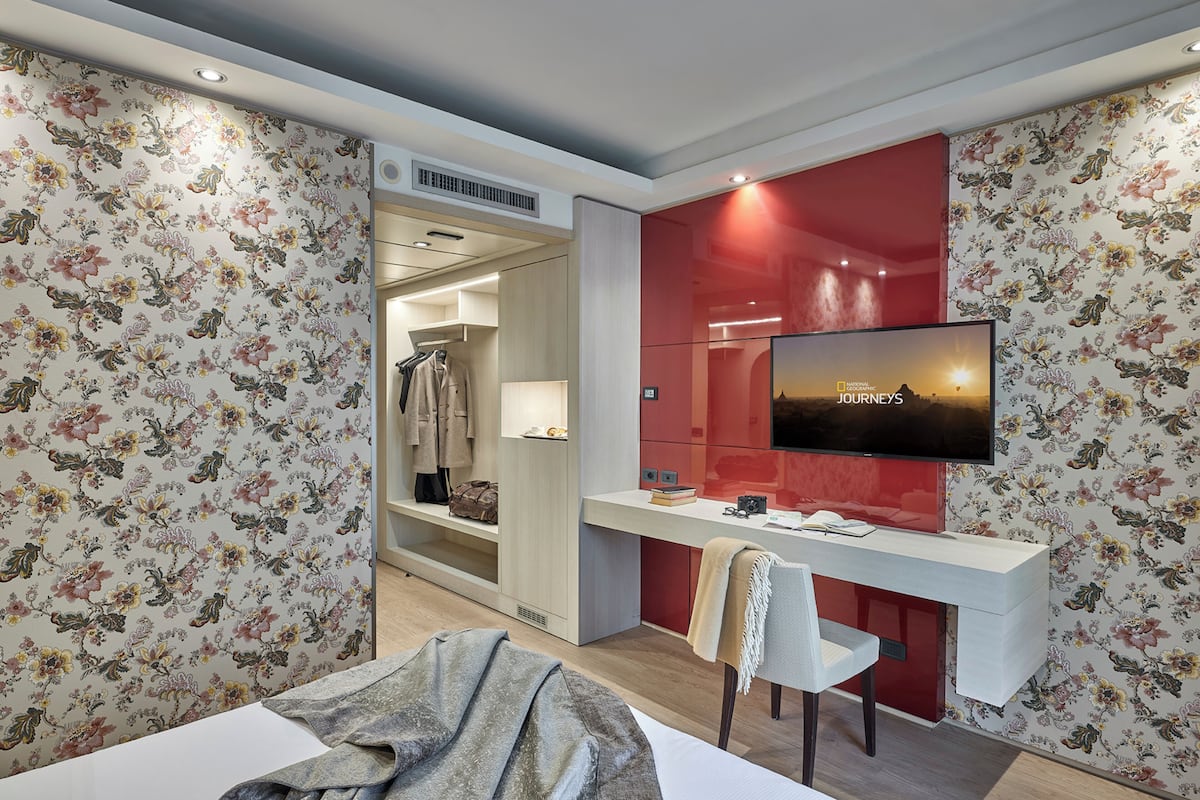 Habitación de hotel con cama, escritorio con silla, televisor montado en la pared y armario.