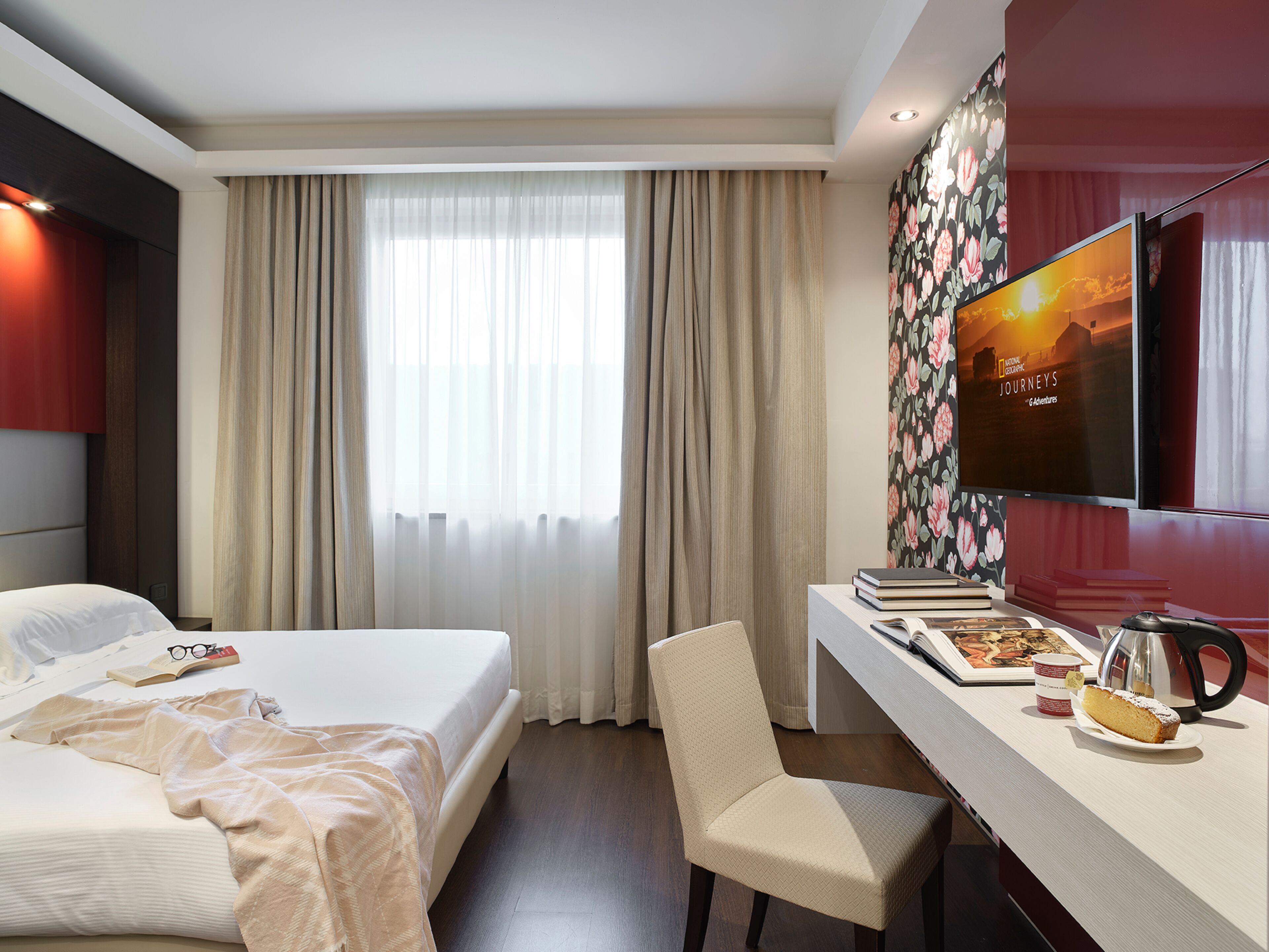 A hotel room with a bed, a desk with a chair, a television, and a window with curtains.