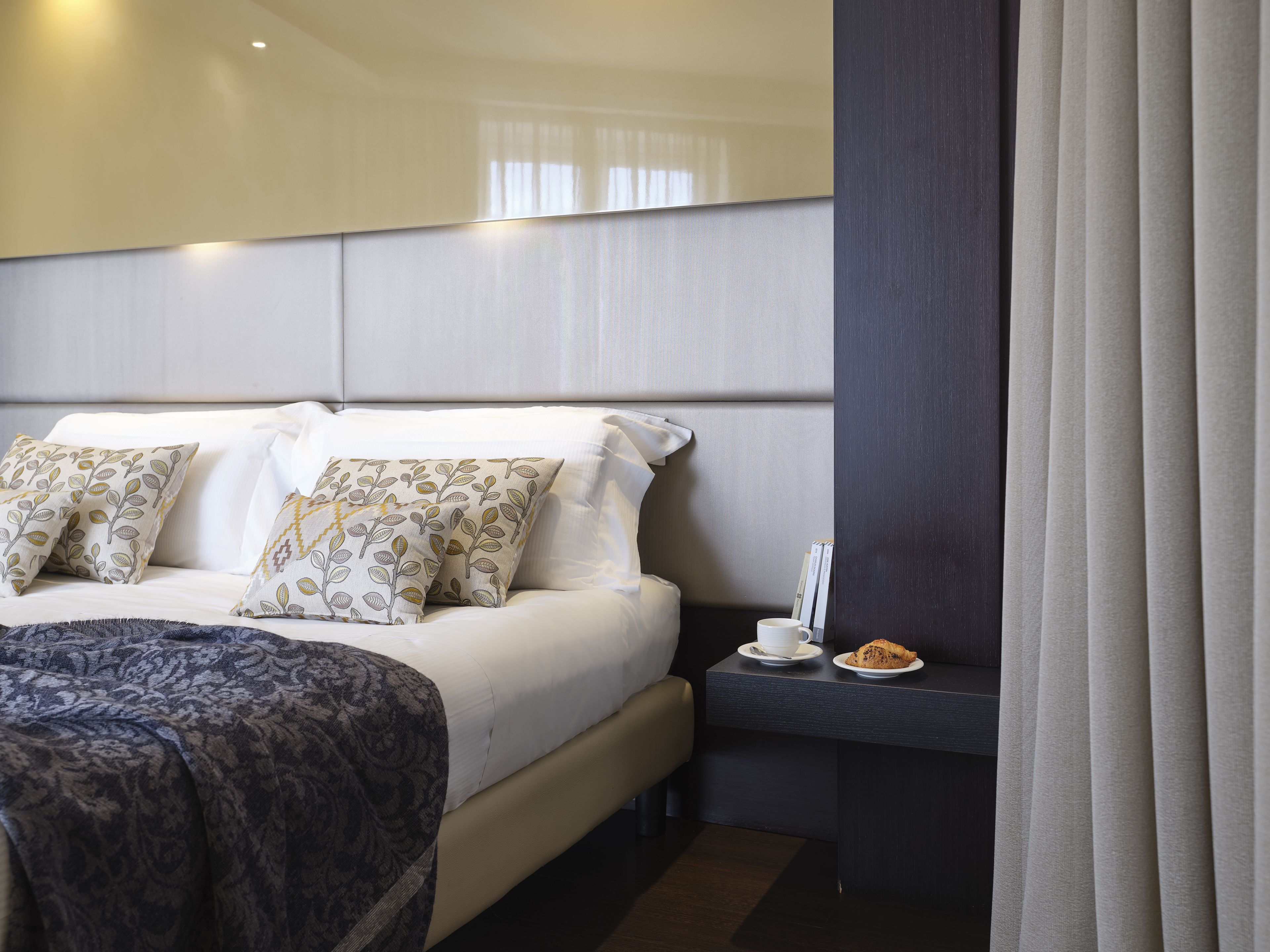 Habitación de hotel con cama, almohadas, una mesita de noche con una taza y un pastel, y una cortina.