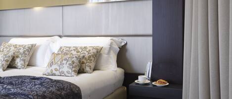 A hotel room with a bed, pillows, a bedside table with a cup and a pastry, and a curtain.