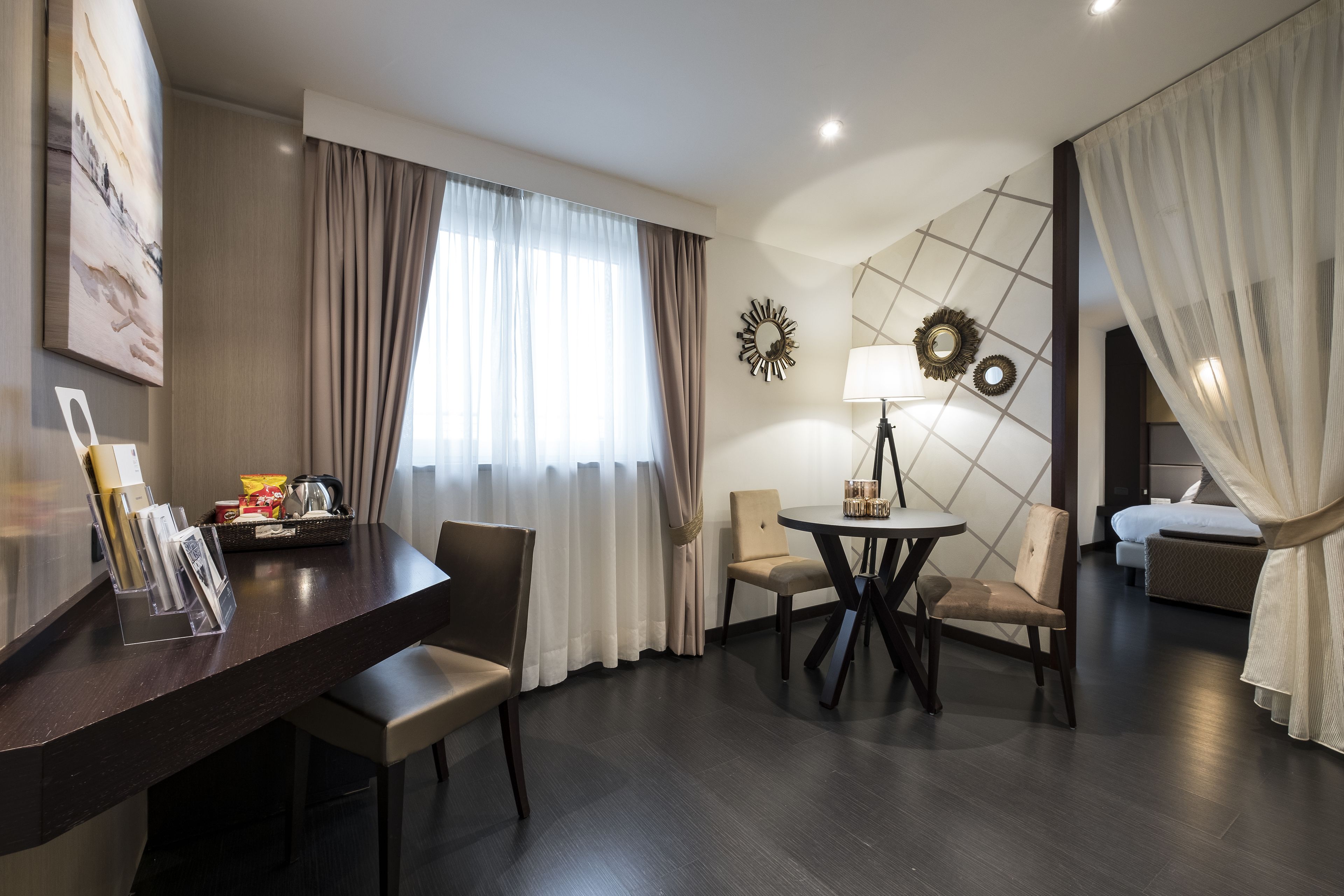 Habitació d'hotel amb sòl de fusta fosca, taula rodona de dinar amb quatre cadires, llit amb roba de llit blanca i una finestra gran amb cortines transparents.