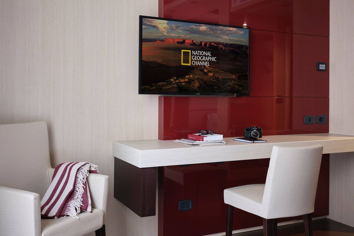 Ein modernes Zimmer mit einem an der Wand befestigten Flachbildfernseher, der den 'National Geographic Channel' zeigt. Es gibt einen Schreibtisch mit Kamera und Büchern, einen Stuhl mit gestreifter Decke und eine rote Kommode.