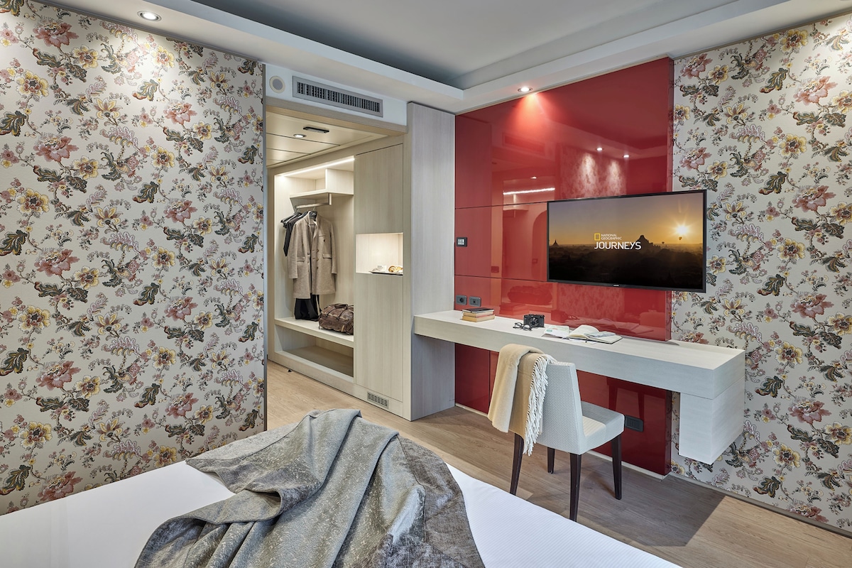 Habitación de hotel con cama, escritorio con silla, televisor montado en la pared y armario.