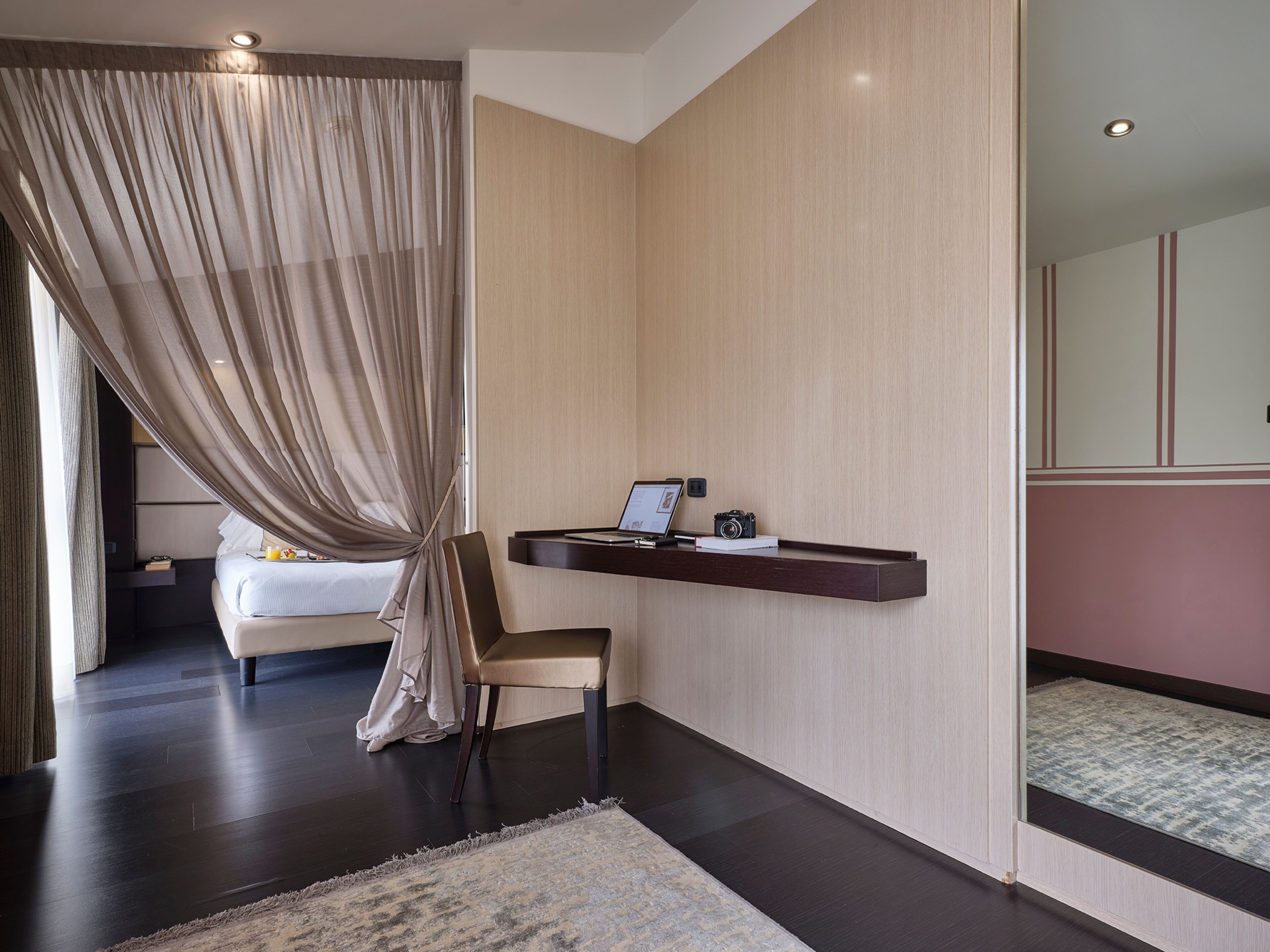 A modern hotel room with a bed, a desk with a laptop and camera, a chair, and a large window with curtains.