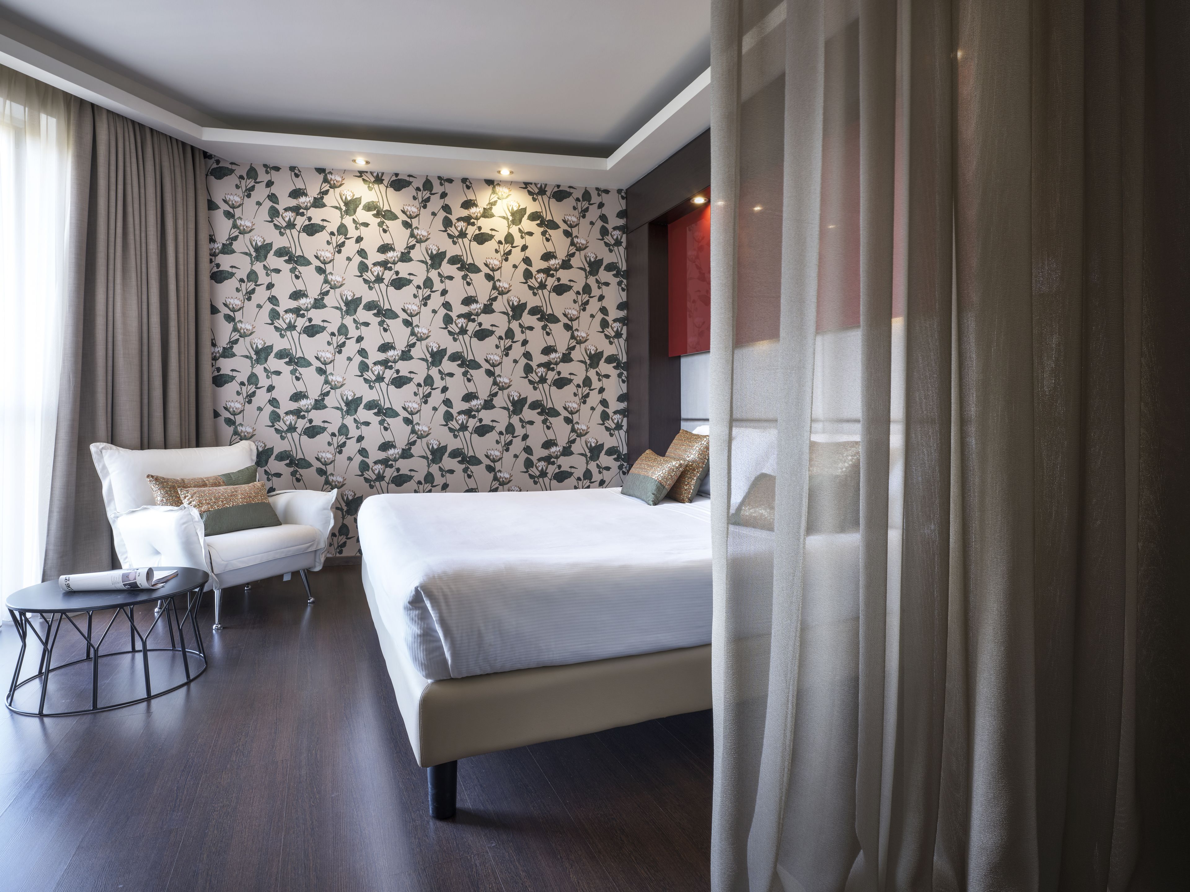 A hotel room with a large bed, a sitting area with a chair and a small table, a decorative wall with a floral pattern, and a window with curtains.