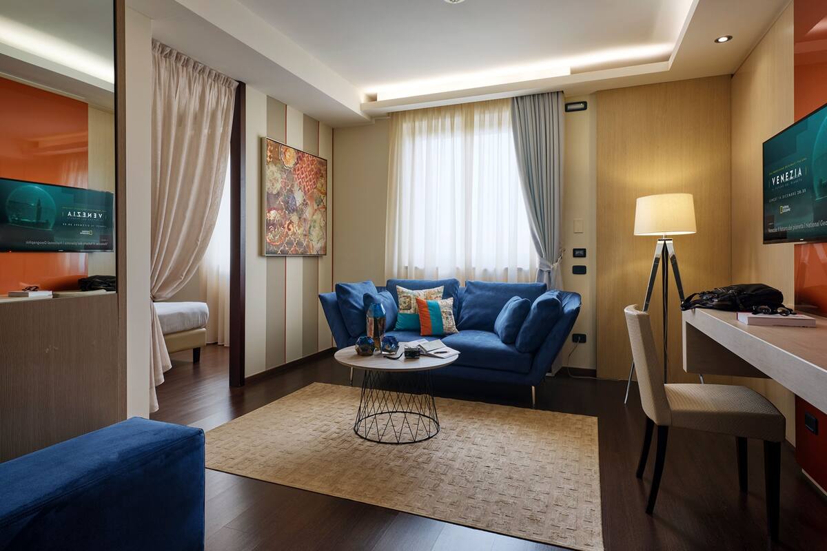 Ein modernes Hotelzimmer mit einem blauen Sofa, einem kleinen Couchtisch, einem Schreibtisch mit Lampe sowie einem an der Wand montierten Fernseher.