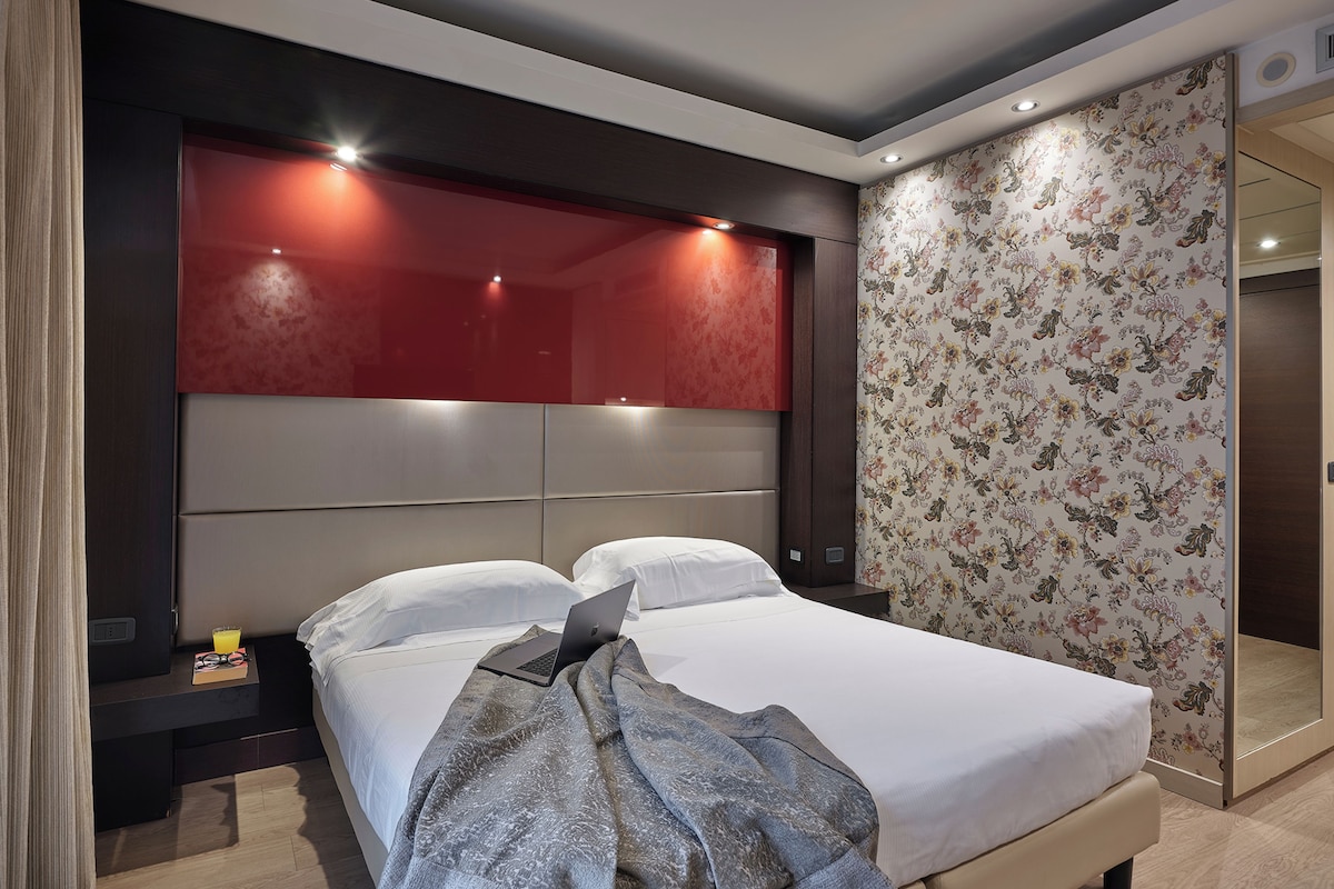 A modern hotel room with a large bed, a nightstand, a laptop, and a red accent wall.