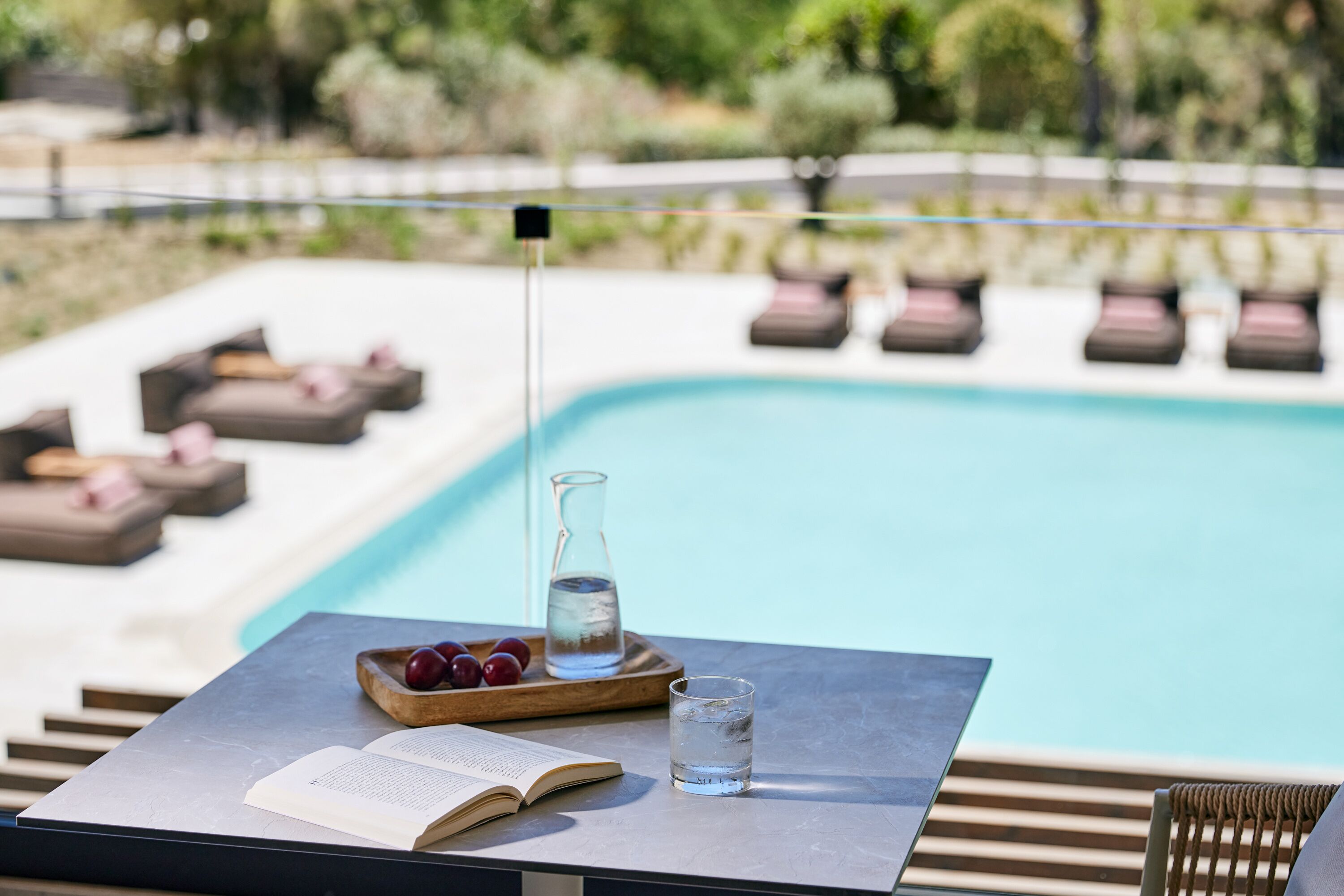 A poolside area with a table, a book, a glass of water, and a bowl of fruit.
