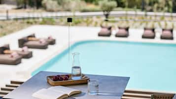 A poolside area with a table, a book, a glass of water, and a bowl of fruit.