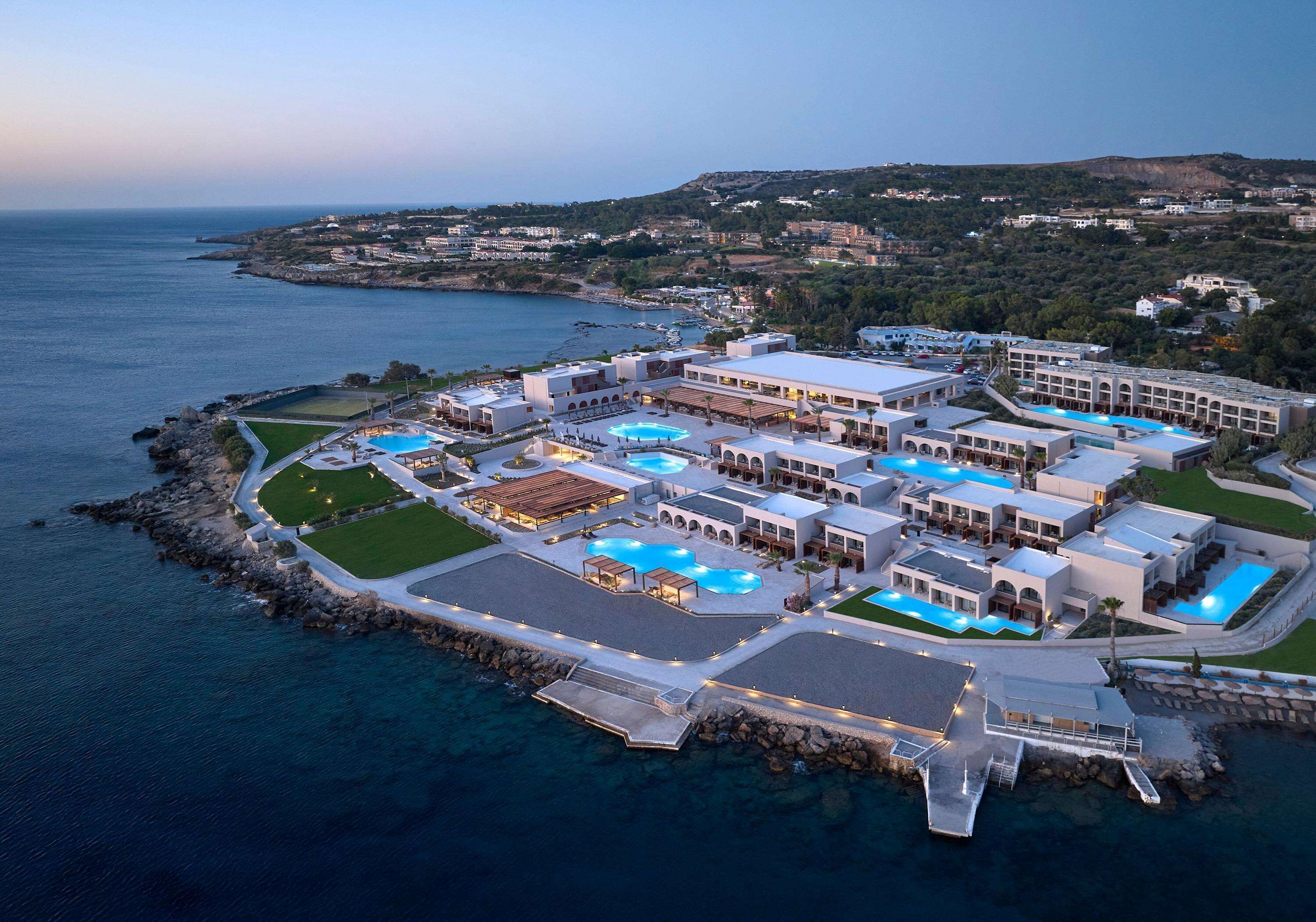 An aerial view of a coastal hotel with multiple swimming pools, a marina, and a rocky shoreline.