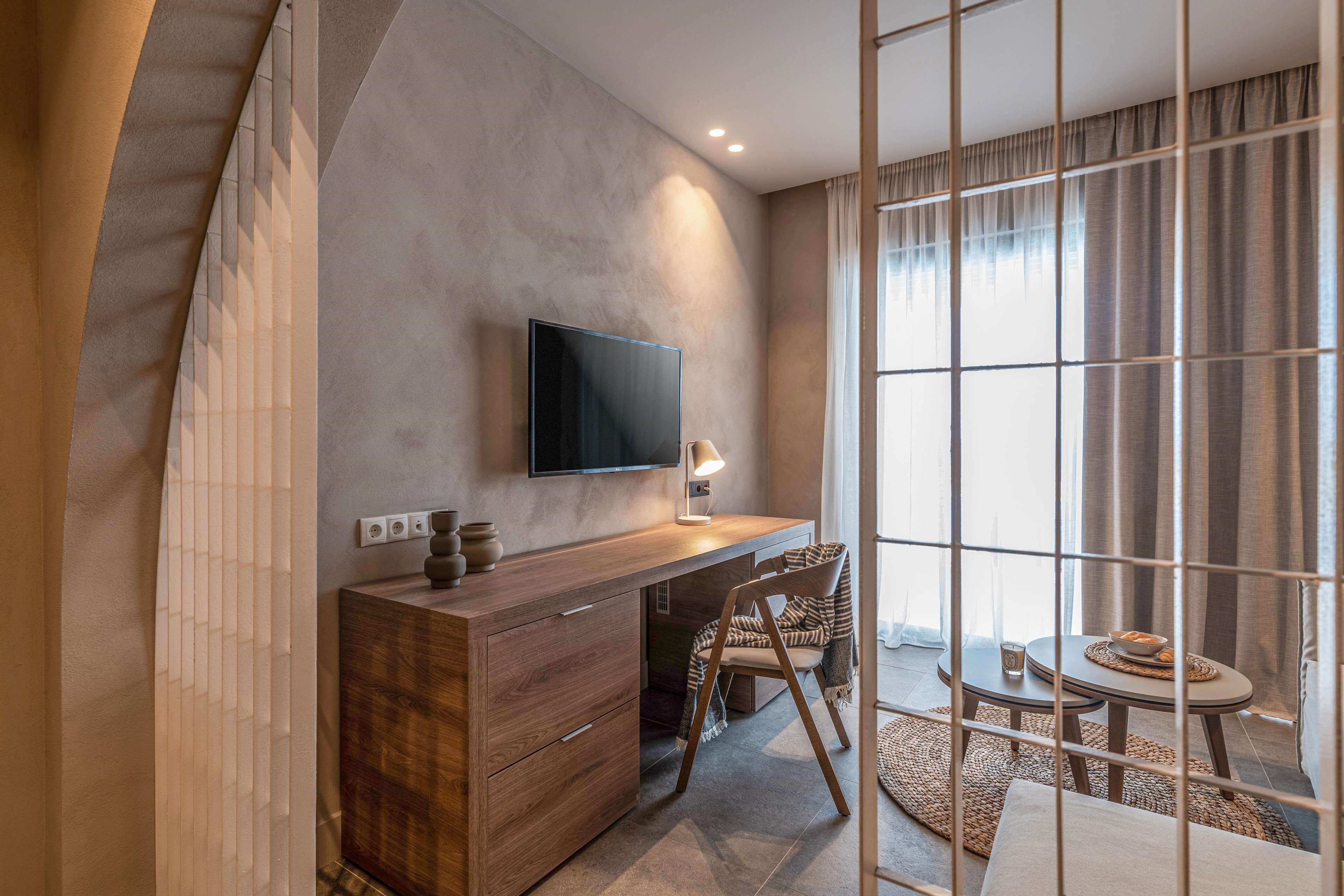 Ein modernes Hotelzimmer mit einem Holztisch, einem Flachbildfernseher an der Wand, einem bequemen Sessel, einem kleinen runden Tisch mit Obstschale und einem großen Fenster mit Vorhängen.