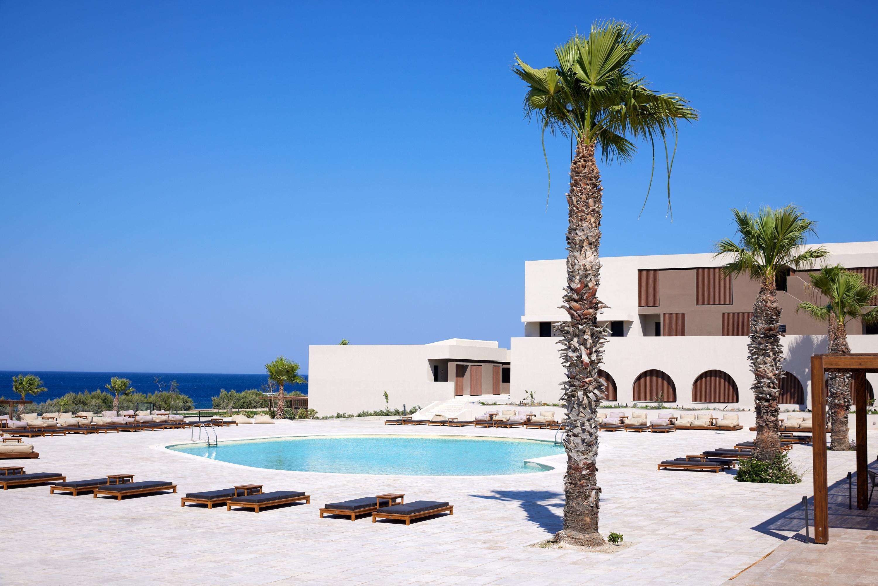 A pool area with lounge chairs and palm trees, a building with arched windows, and a clear blue sky.