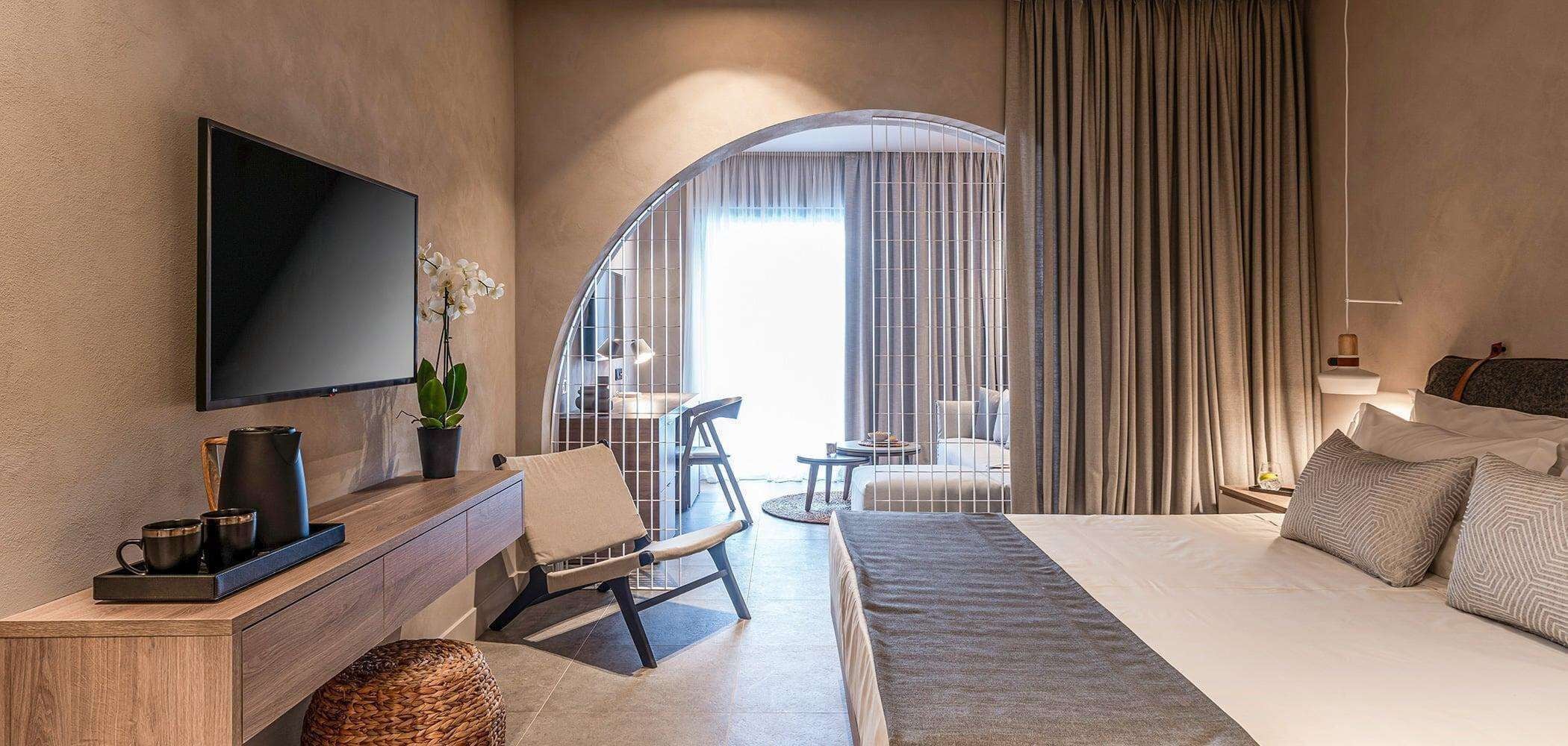 Ein modernes Hotelzimmer mit Flachbildfernseher, einer Sitzecke mit Sessel und Tisch, einem Bett mit Kissen sowie Blick durch ein großes Fenster mit Vorhängen.