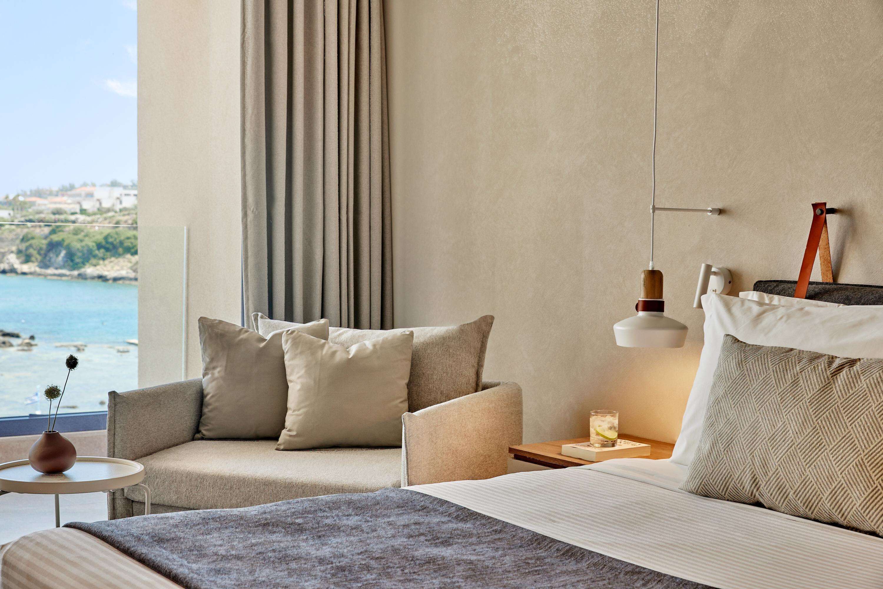 A modern hotel room with a large bed, two armchairs, a small table, and a view of the sea.