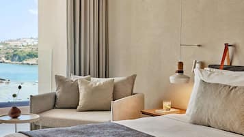 A modern hotel room with a large bed, two armchairs, a small table, and a view of the sea.