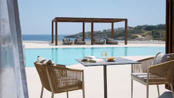 A poolside area with wicker chairs, a table with a book and a bowl of fruit, and a wooden pergola overlooking a beach.