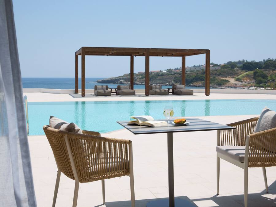 A poolside area with wicker chairs, a table with a book and a bowl of fruit, and a wooden pergola overlooking a beach.