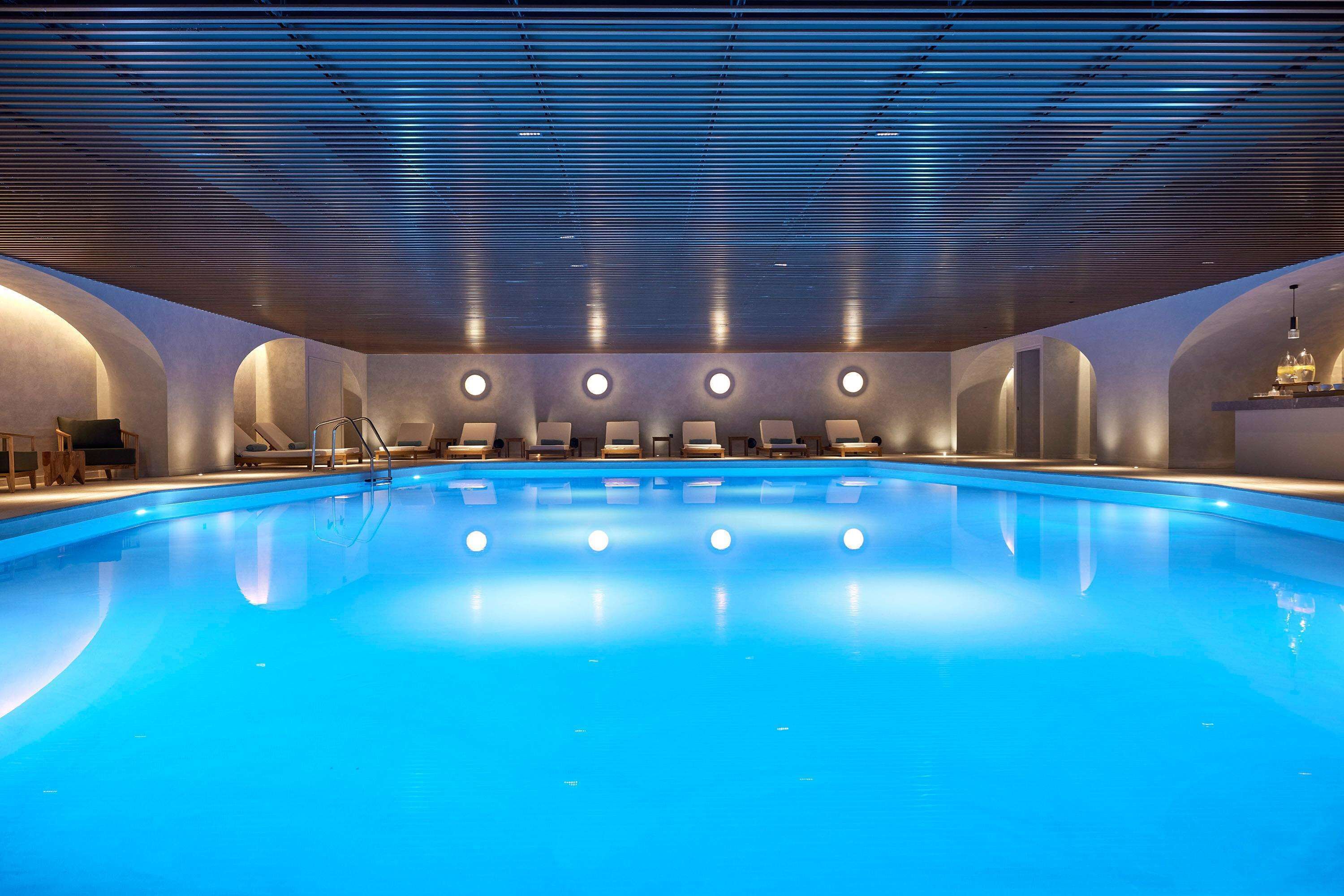 A large indoor swimming pool with a modern design, featuring a blue pool, seating areas with loungers, and a ceiling with horizontal lines.