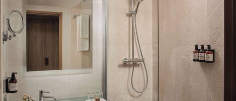 A modern bathroom with a glass sink, a large mirror, a shower with a handheld showerhead, and toiletries on the wall.