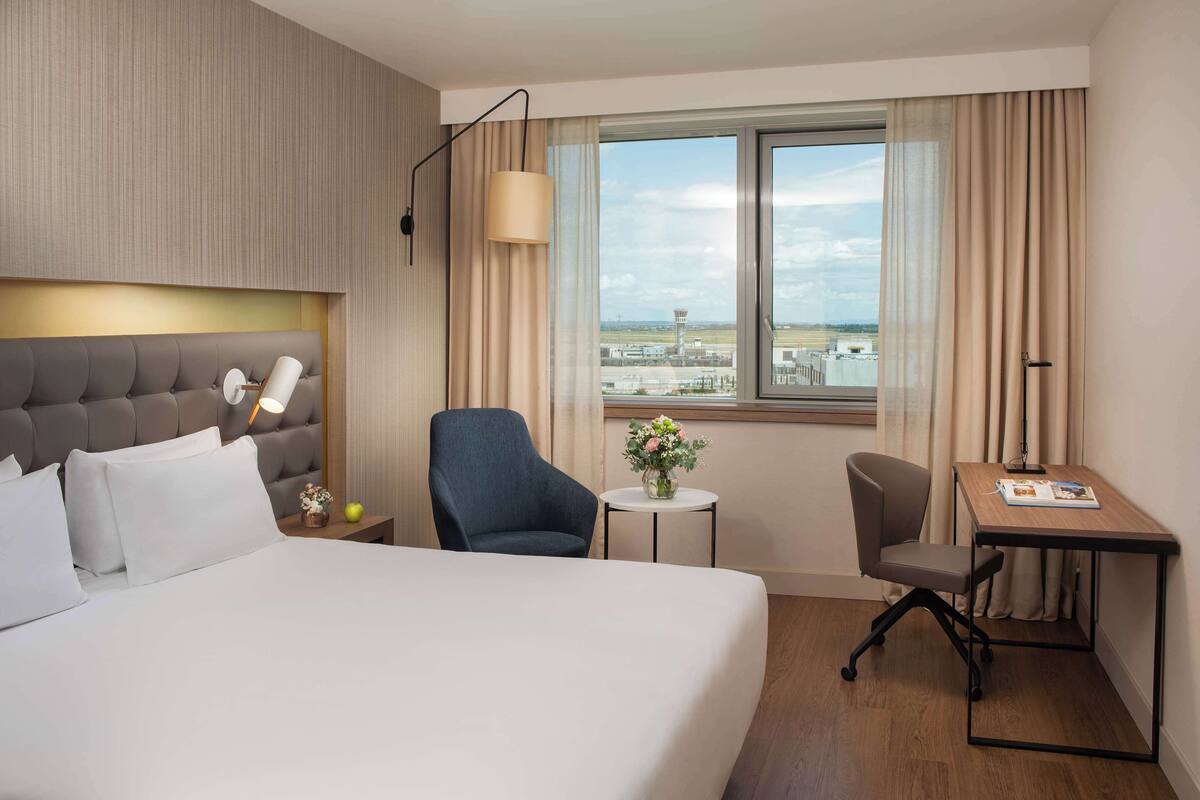 Ein Hotelzimmer mit einem großen Bett, einem Schreibtisch, einem Stuhl, einer Lampe, einer Vase mit Blumen und Blick auf einen Flughafen.