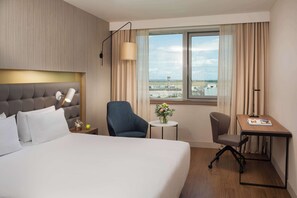 A hotel room with a large bed, a desk, a chair, a lamp, a vase with flowers, and a view of an airport.