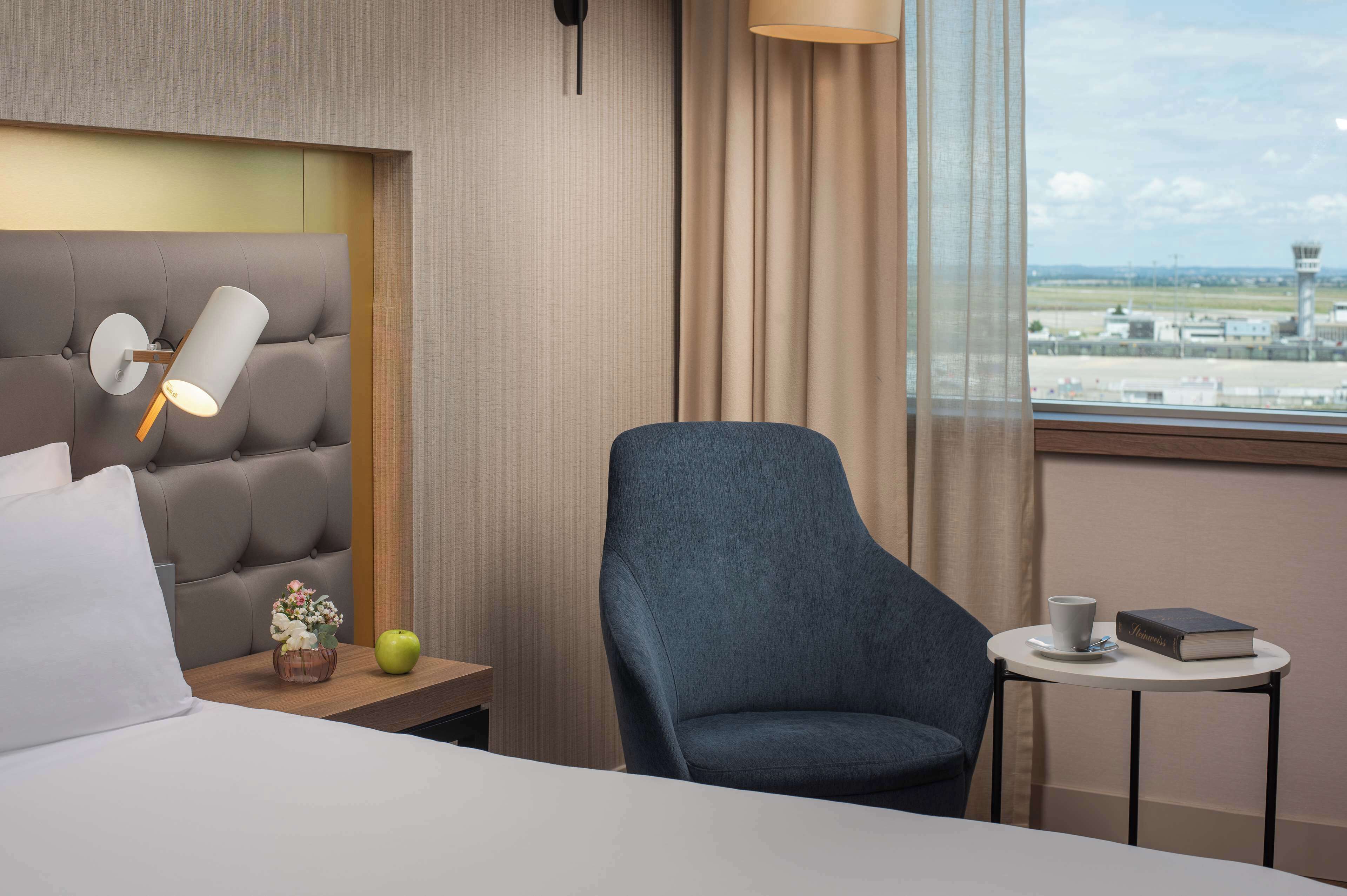 Hotelkamer met een bed, nachtkastje met een lamp, een stoel, een klein tafeltje met een boek en een kopje, en een raam met uitzicht op een vliegveld.