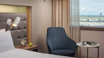 Hotelkamer met een bed, nachtkastje met een lamp, een stoel, een klein tafeltje met een boek en een kopje, en een raam met uitzicht op een vliegveld.