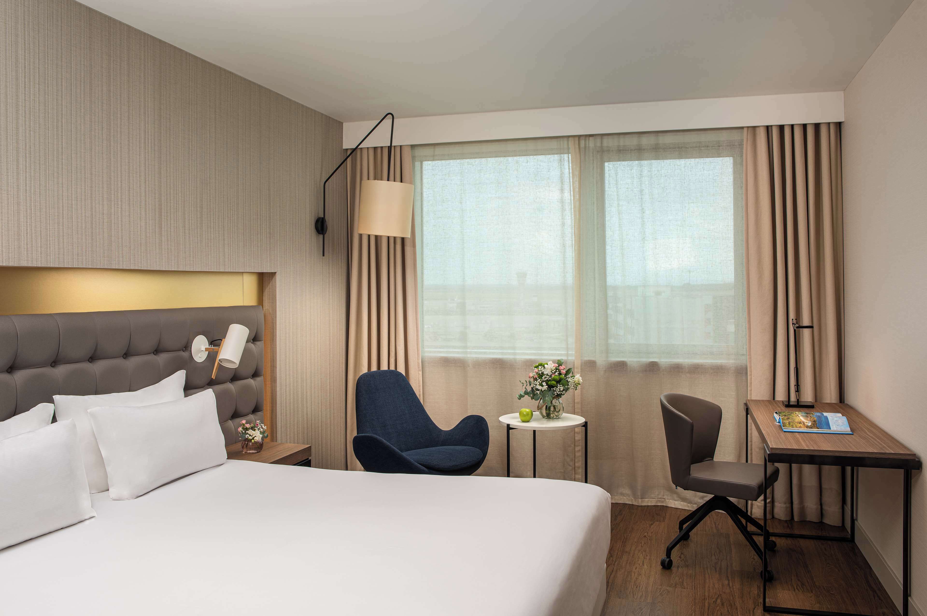 A hotel room with a large bed, a desk, two chairs, a small table with a vase of flowers, and a window with curtains.