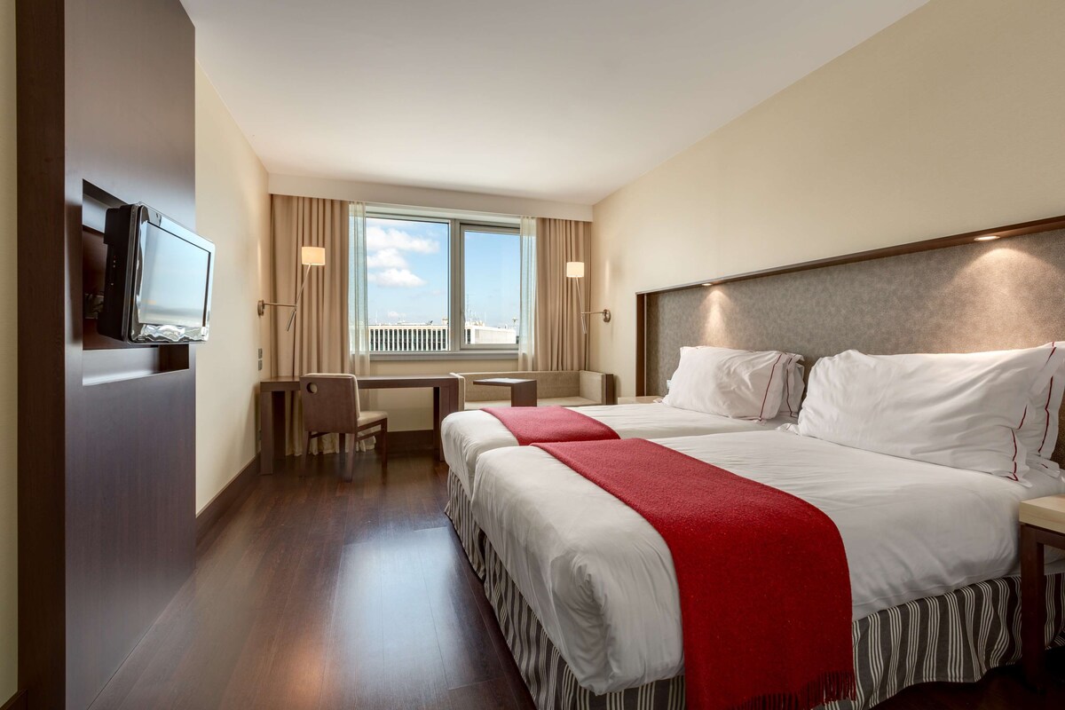 A hotel room with a bed, a desk, a chair, a TV, and a window with a view.