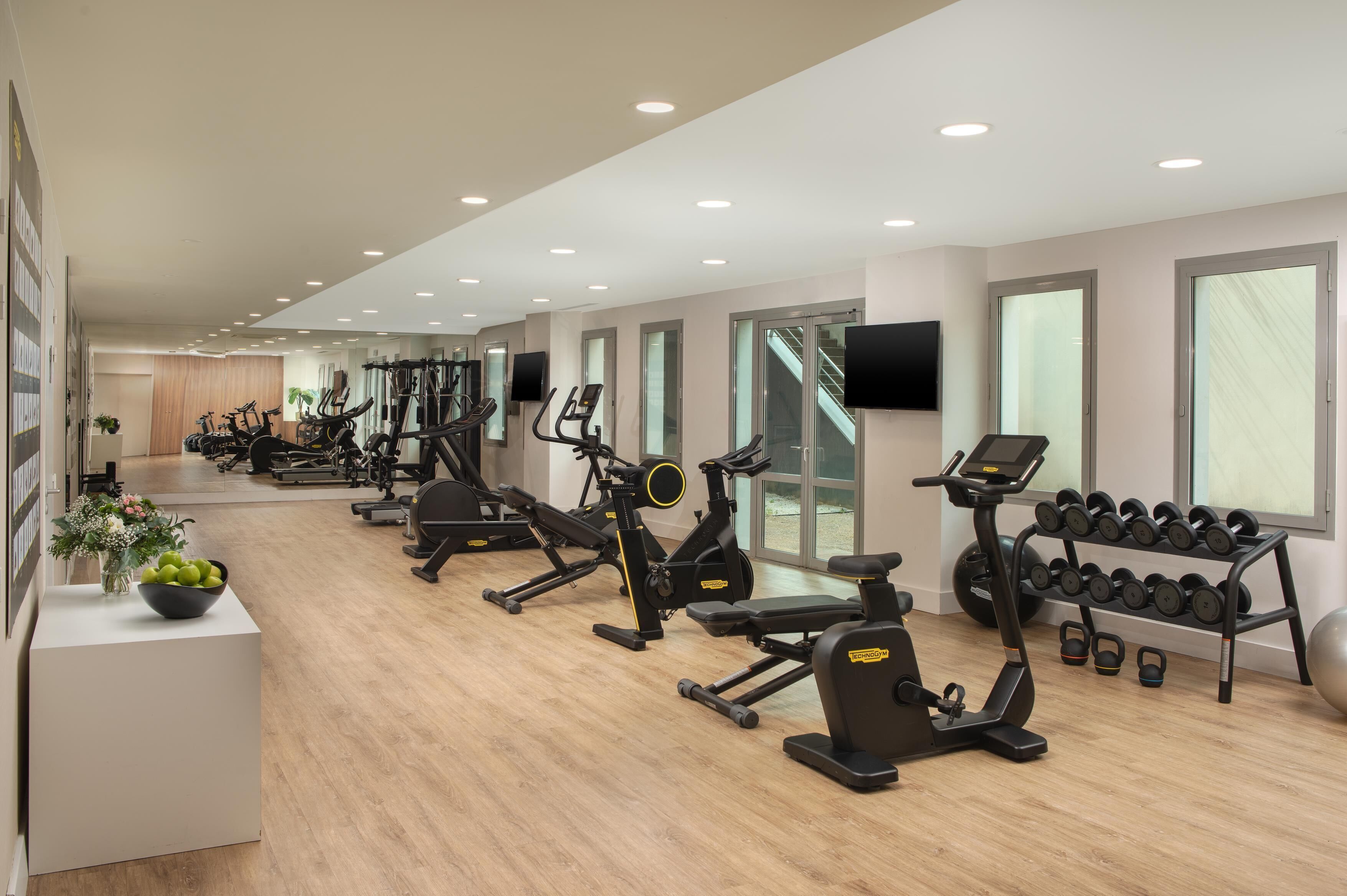 A gym with various exercise machines, a wall-mounted TV, and a rack of dumbbells.