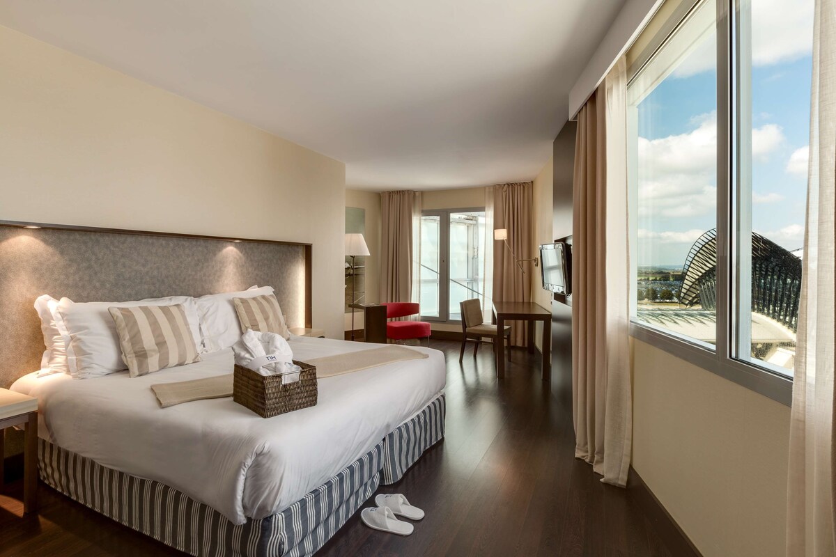 A hotel room with a large bed, a desk, a chair, a TV, and a view of the cityscape.