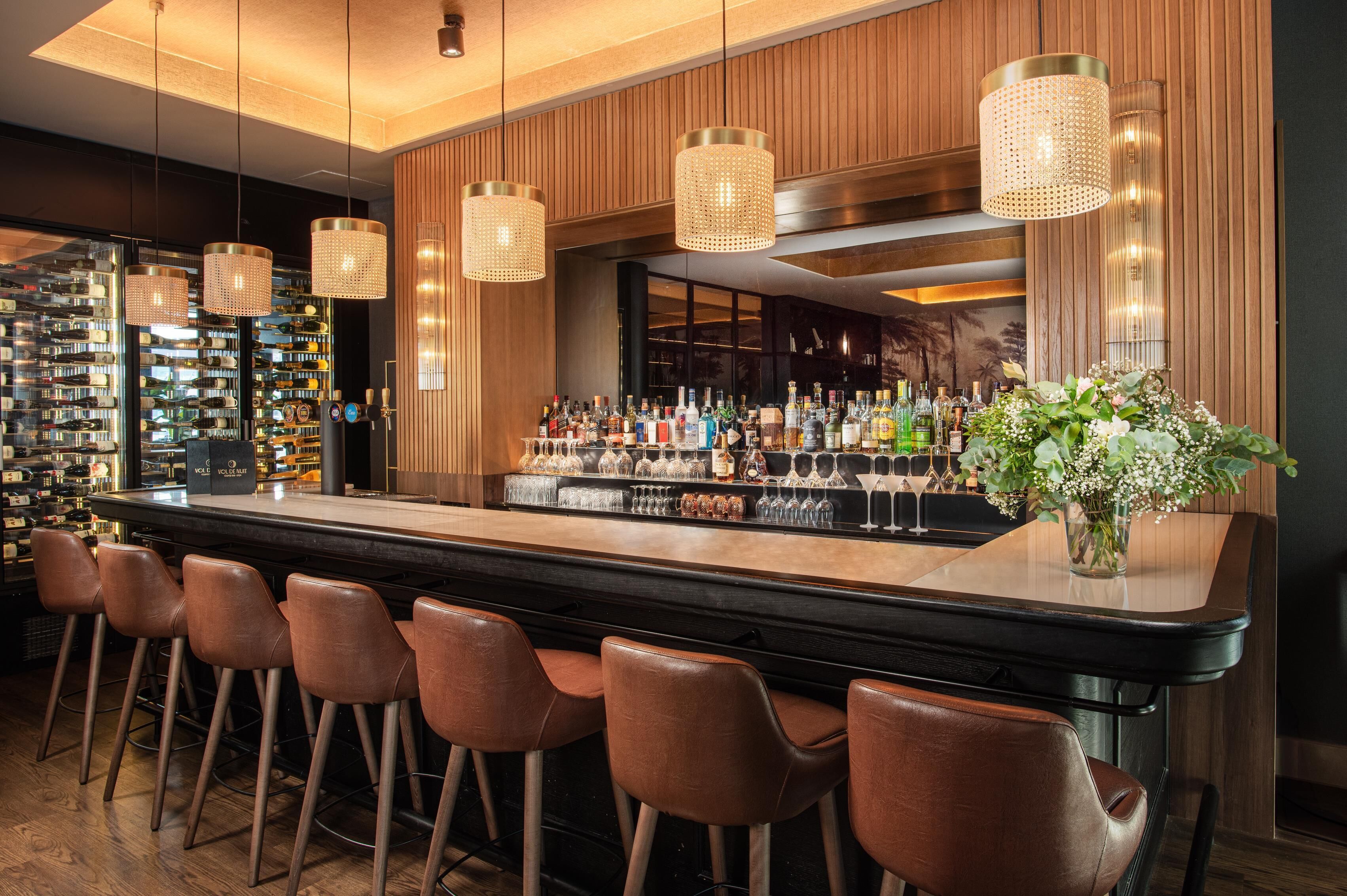 A bar area with a long counter, multiple bottles, glasses, and a decorative plant.