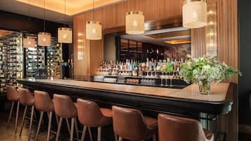 Una zona de bar con una barra larga, varias botellas, vasos y una planta decorativa.