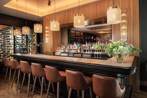 A bar area with a long counter, multiple bottles, glasses, and a decorative plant. - NH Lyon Airport (Colombier-Saugnieu)