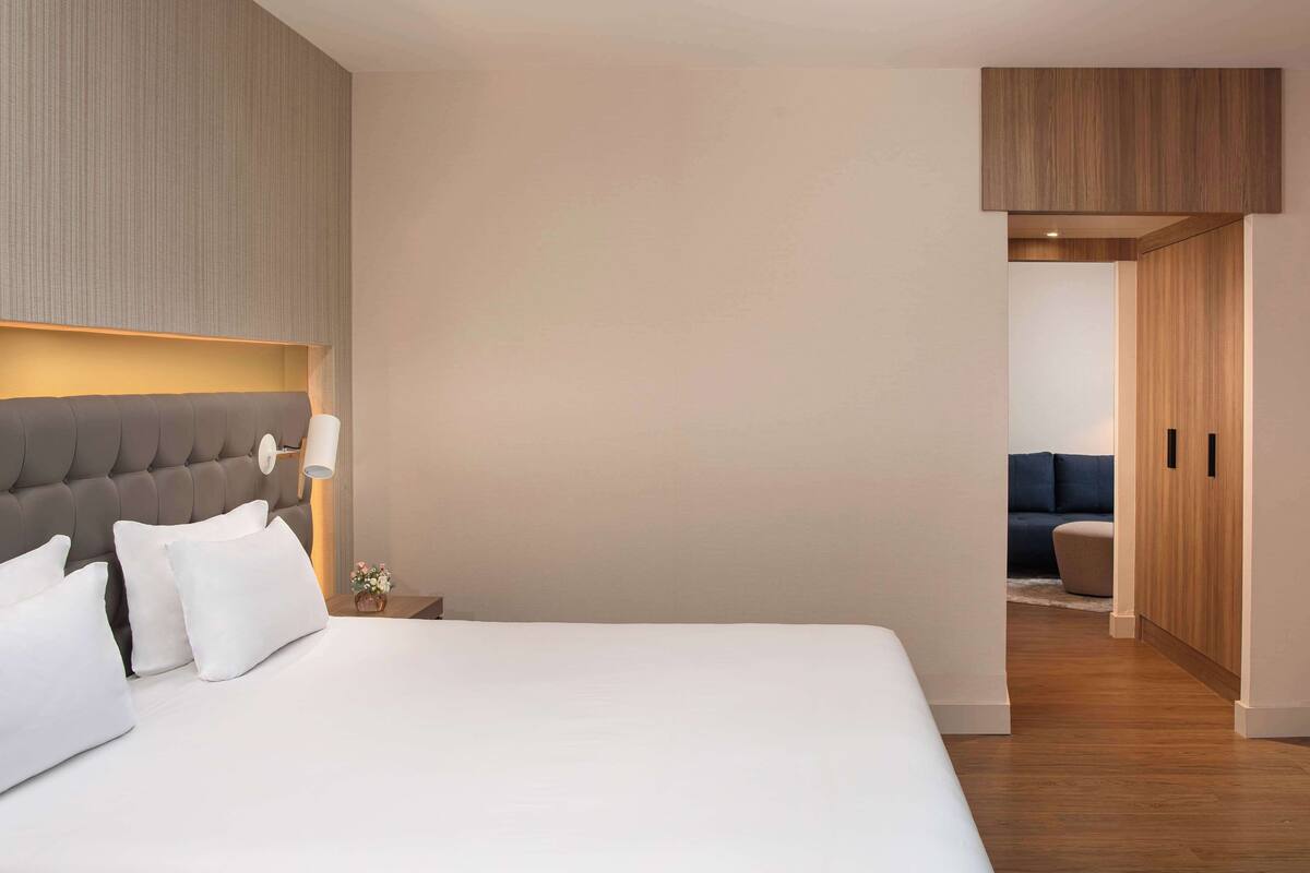 Ein modernes Hotelzimmer mit einem großen Bett, einem Nachttisch, einer Lampe und Blick in einen anderen Raum mit einer Couch.