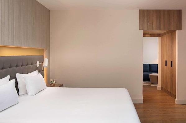 A modern hotel room with a large bed, a nightstand, a lamp, and a view into another room with a sofa.